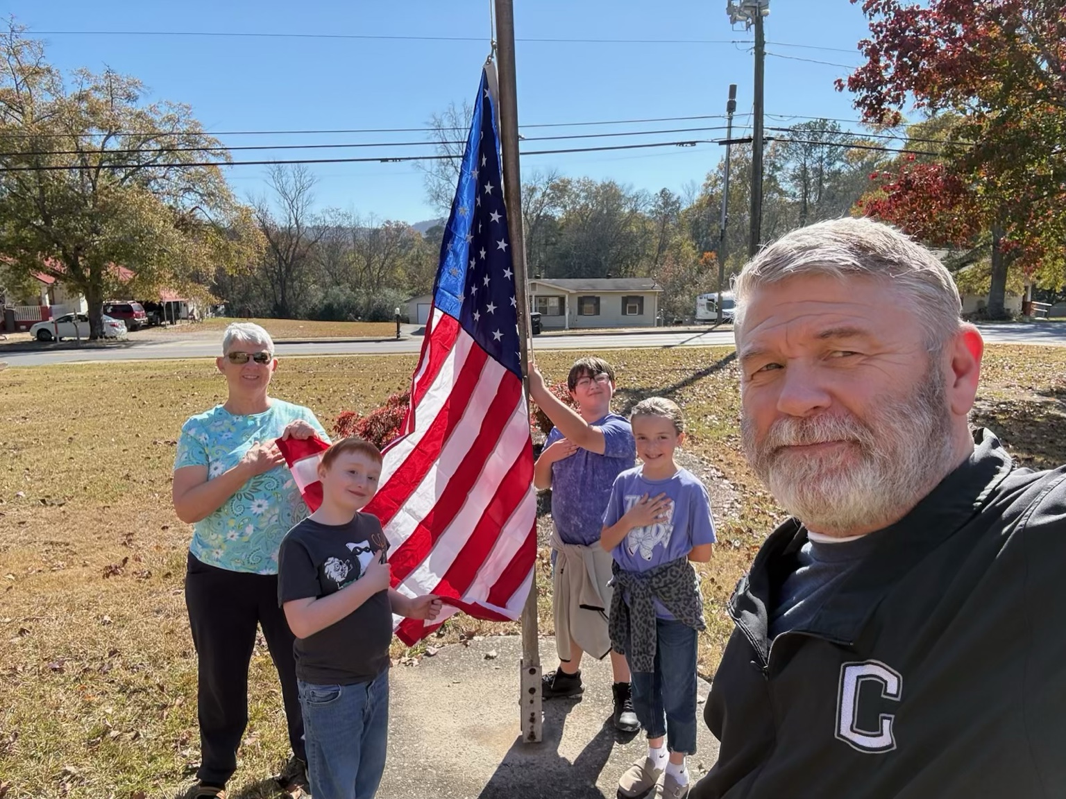 Two adults and three children stand outdoors beside an American flag and flagpole.