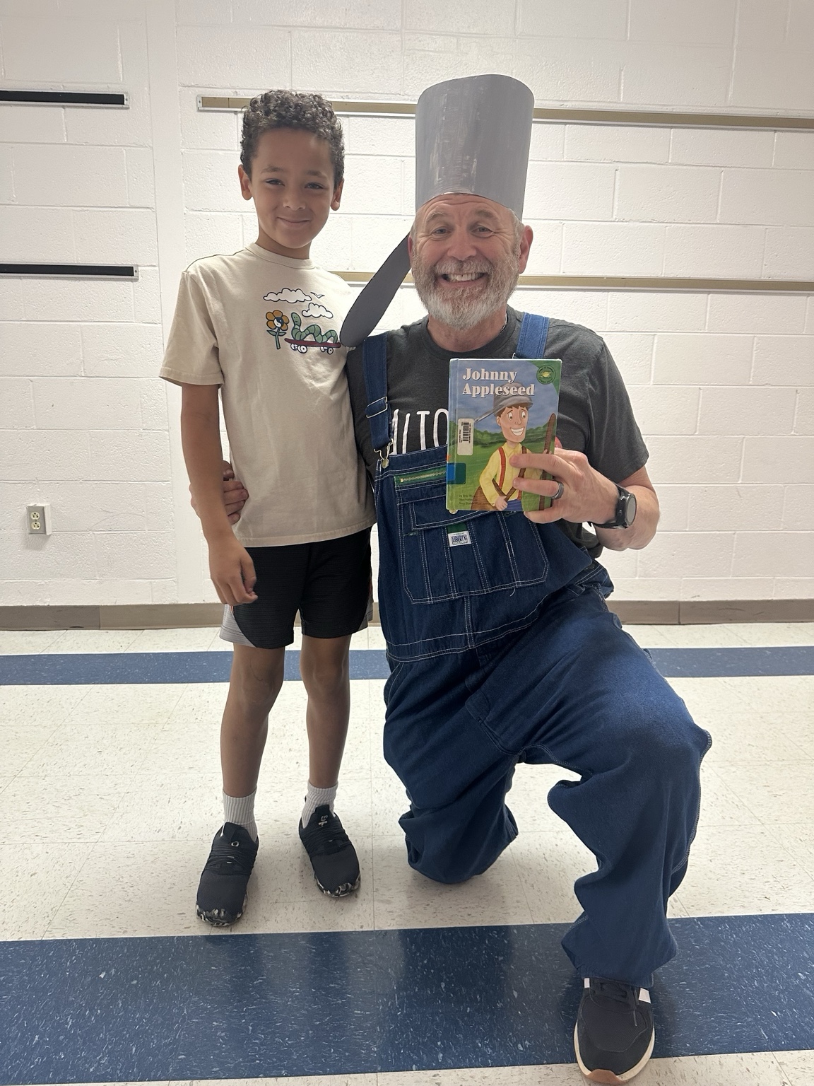 An adult wearing overalls and a tall costume hat holds a Johnny Appleseed book while posing beside a child in a school hallway.