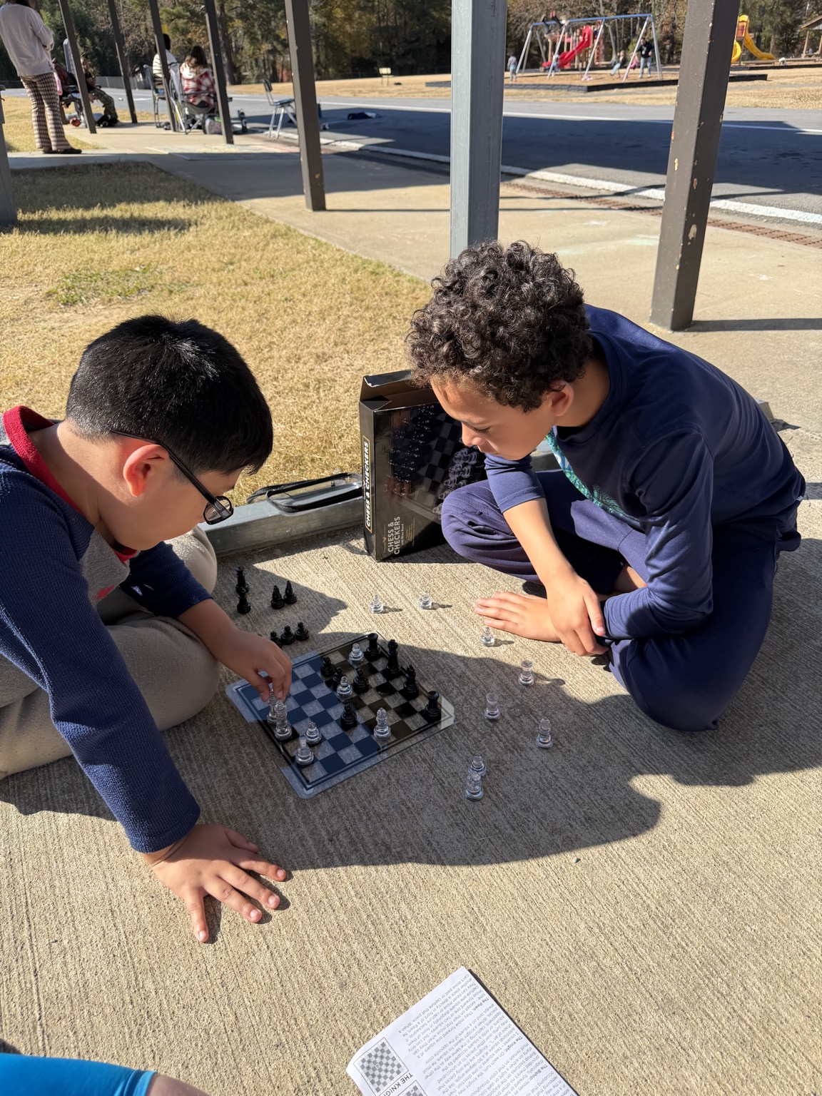 Two children concentrate on a game of chess outdoors beside a playground.
